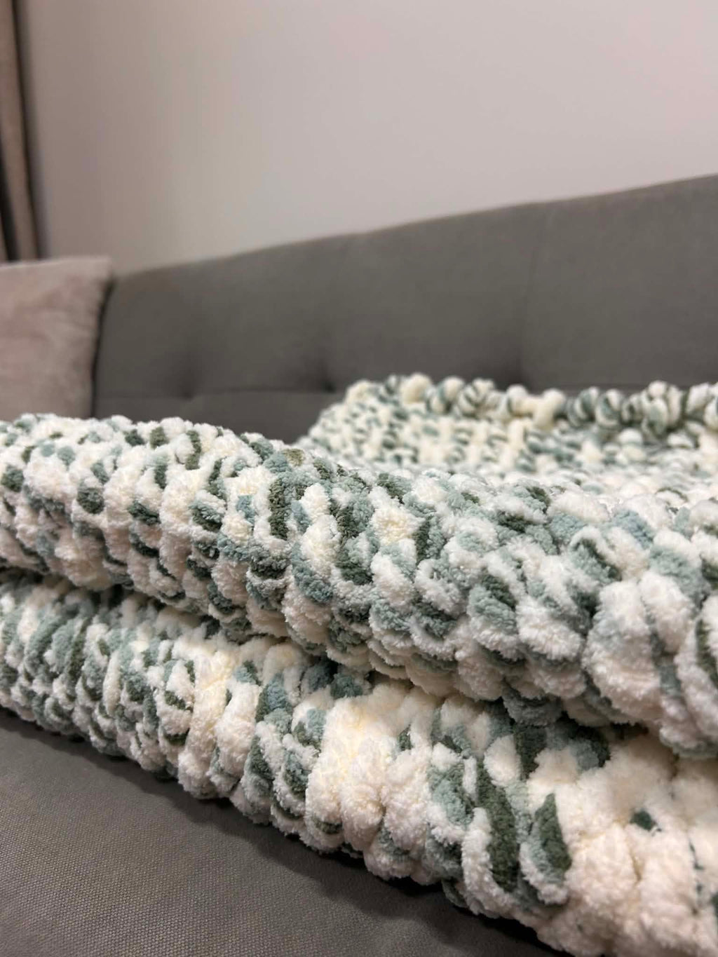 Handmade chunky knit blanket in sage green and cream, folded on a grey sofa